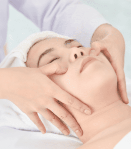 A person with their eyes closed is lying down and receiving a facial massage. Towels cover their hair, and the hands of the masseuse are gently pressing on the person's cheeks and jawline. The scene exudes relaxation, akin to the tranquility found in Acupuncture Long Island sessions.