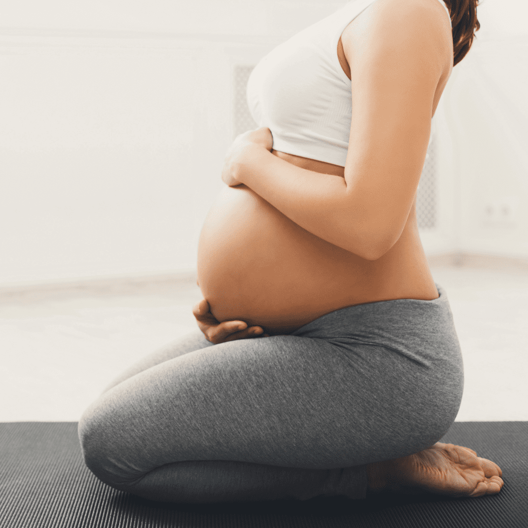 A pregnant person kneels on a yoga mat, cradling their belly with both hands. They are dressed in a white sports bra and gray leggings, surrounded by the calm atmosphere of a softly lit room. This serene moment highlights the holistic approach of Fertility Treatments Long Island.