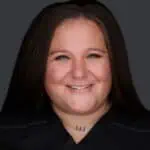 A person with long brown hair and a friendly smile is posing for a portrait against a dark background. They are wearing a dark top and have a small, visible tattoo on their neck below the collarbone, reflecting their journey through pain management on Long Island.