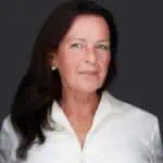 A woman with long dark hair is wearing a white blouse and small earrings. She is standing against a dark gray background, looking directly at the camera with a slight smile, as if she's just experienced the benefits of acupuncture on Long Island.