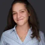 A woman with long brown hair is smiling softly at the camera. She is wearing a blue and white striped button-up shirt and a necklace with a small pendant. The background, fitting for an Acupuncture Long Island studio, is solid dark gray.