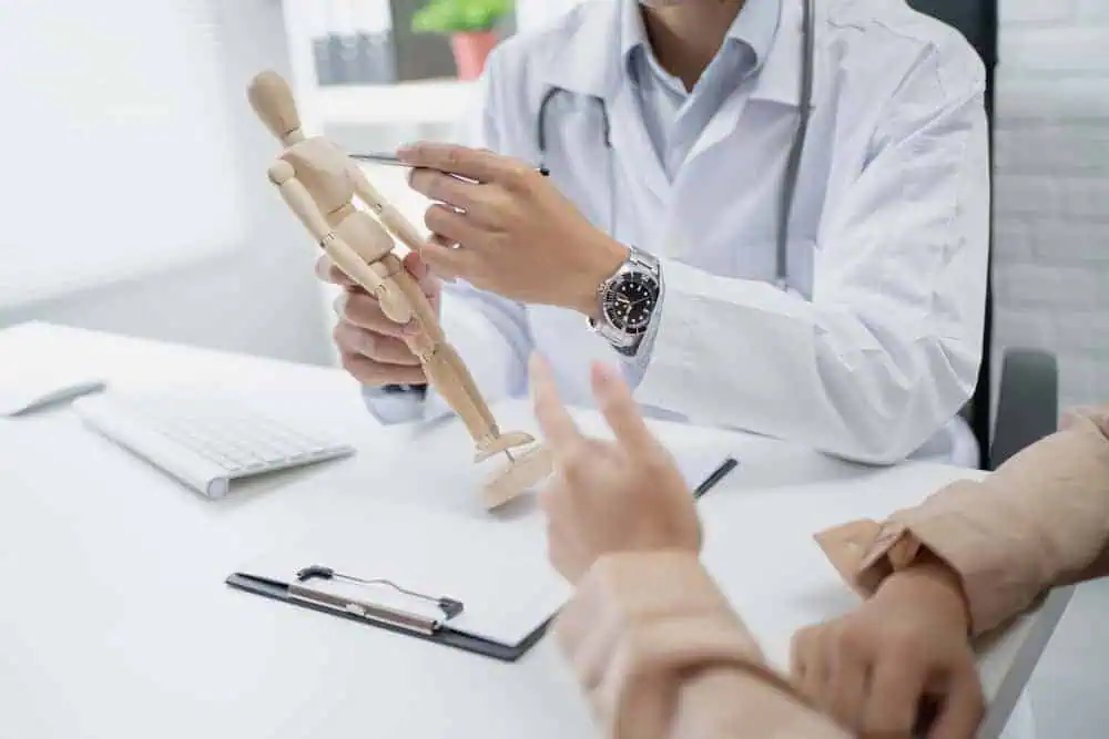 A doctor in a white coat is holding a wooden anatomical figure, discussing acupuncture with a patient across the table. On the table are a keyboard and a clipboard with papers detailing Pain Management Long Island options.