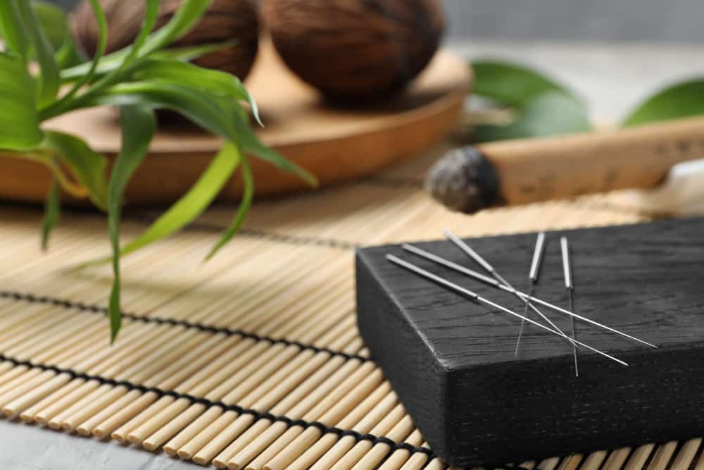 A close-up view of acupuncture needles resting on a black wooden block. The background features a bamboo mat, a green plant, and a rustic wooden bowl with round objects, creating a serene and natural ambiance reminiscent of Acupuncture Long Island’s holistic approach.