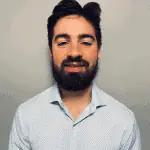 A man with dark hair and a full beard is smiling at the camera. He is wearing a light blue button-up shirt with a subtle checkered pattern, standing against a plain gray background. His expertise in Acupuncture Long Island brings balance and well-being to his clients.