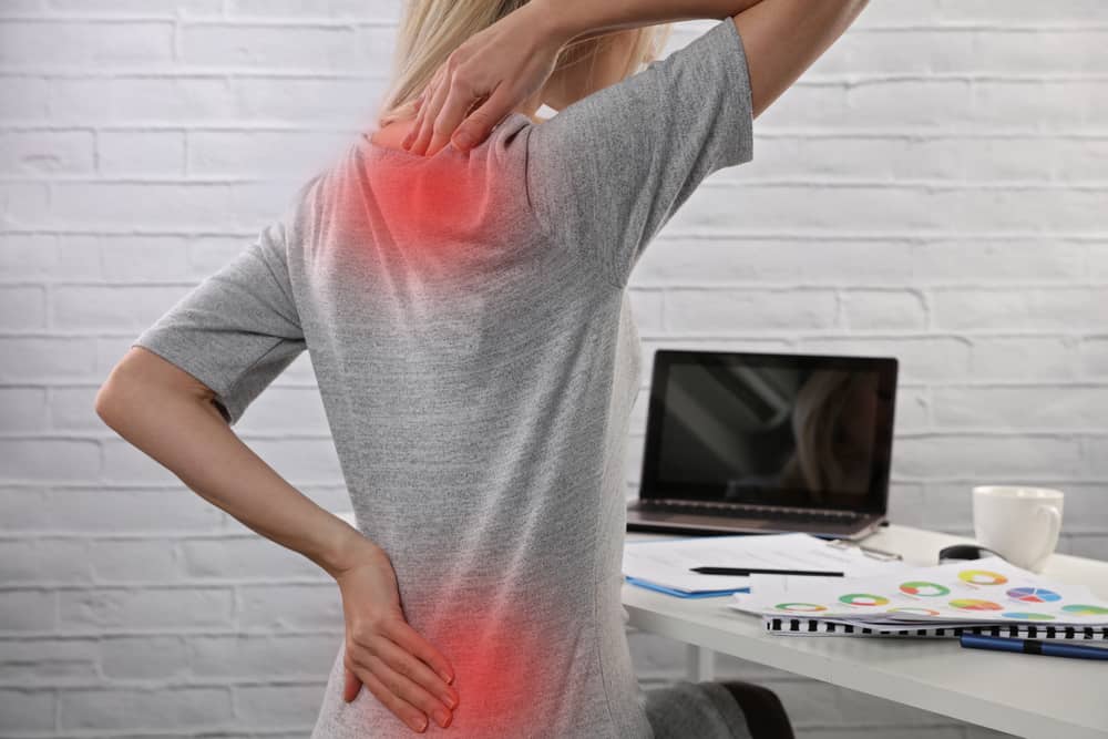 A person wearing a gray shirt is sitting at a desk, touching their upper and lower back where red-colored areas indicate pain. The desk has an open laptop, papers, a notebook, and a white mug. The background features a white brick wall—a perfect setting for exploring Pain Management Long Island solutions.