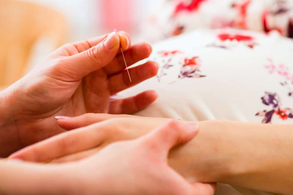 A person performing acupuncture on another person's hand. The hand is resting as a thin needle is inserted into the skin. The background features a floral pattern, possibly on clothing.
