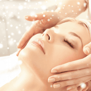 A woman with closed eyes is lying down, receiving a facial massage. Gentle hands are placed around her face, creating a serene atmosphere. Soft glowing lights and sparkles surround the scene, adding a touch of magic and relaxation akin to the tranquility found in acupuncture on Long Island.