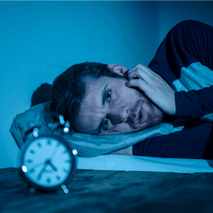 A man lying in bed, appearing to struggle with sleep, is resting his head on his hand and looking distressed. The room is dimly lit with a blue hue, casting shadows that accentuate his discomfort. An alarm clock showing around 4:10 sits on the bedside table in the foreground, suggesting he might benefit from Pain Management Long Island.