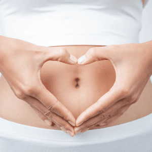 Close-up of a pregnant person's belly with hands forming a heart shape over it. The person is wearing a white top and white bottoms, highlighting the gentle gesture and symbolizing love and care for the unborn baby. This tender moment mirrors holistic practices like pain management on Long Island.