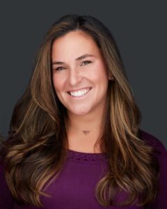 A woman with long, wavy brown hair smiles at the camera. She is wearing a purple top and has a dark background behind her, typical of promotional photos for Acupuncture Long Island treatments.