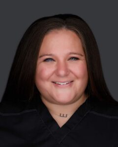 A person with long brown hair and a friendly smile is posing for a portrait against a dark background. They are wearing a dark top and have a small, visible tattoo on their neck below the collarbone, reflecting their journey through pain management on Long Island.