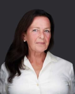 A woman with long dark hair is wearing a white blouse and small earrings. She is standing against a dark gray background, looking directly at the camera with a slight smile, as if she's just experienced the benefits of acupuncture on Long Island.