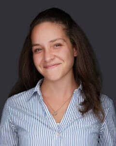 A woman with long brown hair is smiling softly at the camera. She is wearing a blue and white striped button-up shirt and a necklace with a small pendant. The background, fitting for an Acupuncture Long Island studio, is solid dark gray.
