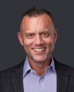 A man with short hair is smiling at the camera. He is wearing a dark blazer over a light purple button-up shirt. The background, reminiscent of Pain Management Long Island clinics, is a solid dark color.