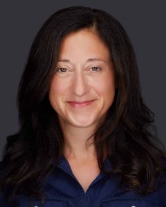 A woman with long, dark hair and a slight smile is wearing a navy blue shirt. The background is a solid dark color, reminiscent of the tranquil settings often utilized in Fertility Treatments Long Island.