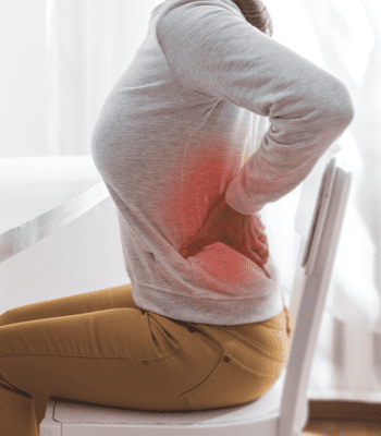 A person sits on a chair, wearing a light grey long-sleeve shirt and mustard-colored pants, with their right hand placed on their lower back. A red-highlighted area on their lower back indicates discomfort, suggesting they might benefit from acupuncture Long Island services.