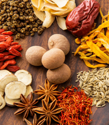 A variety of dried spices and herbs are arranged on a wooden surface. The assortment includes star anise, goji berries, dried red dates, dried ginger slices, whole nutmegs, and dried orange peel, all creating a colorful and textured display—perfect for enthusiasts of Herbology Long Island.