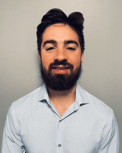 A man with dark hair and a full beard is smiling at the camera. He is wearing a light blue button-up shirt with a subtle checkered pattern, standing against a plain gray background. His expertise in Acupuncture Long Island brings balance and well-being to his clients.